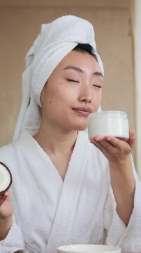 A woman in a white robe and towel smells a jar of cream while holding a coconut half, surrounded by natural ingredients for beauty treatments