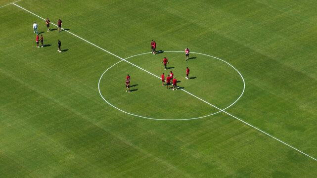 High-angle aerial view of a green soccer field. A group of players in red uniforms is gathered in the center circle for a training session under bright sunlight. Sports concept.