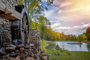 An old wooden water wheel attached to a stone wall turns under flowing water next to a peaceful lake surrounded by lush green forest.