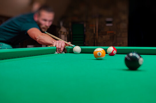 A focused man in a dimly lit pool hall leans over a green felt table to take a precise shot with his cue stick.