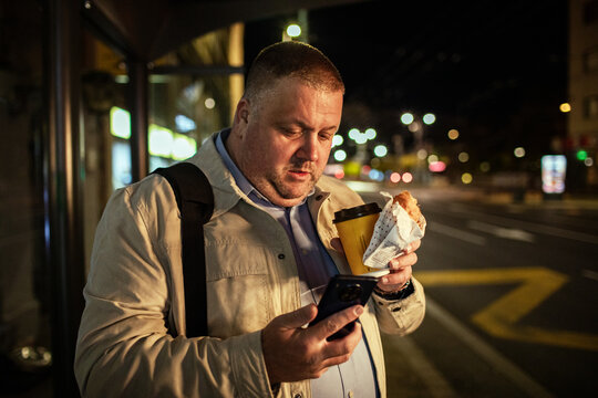 Man checking phone while holding coffee and pastry at night bus stop