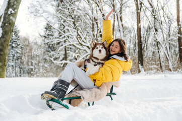 Obraz premium Young woman in yellow winter jacket sitting on a sled with a dog in snowy forest, surrounded by trees and snow-covered ground during a sunny winter day