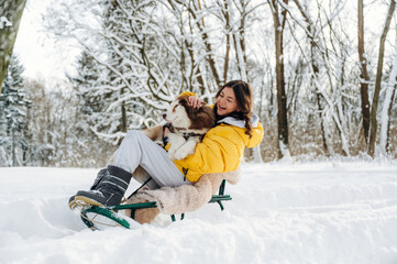 Obraz premium Young woman with long dark hair wearing a yellow jacket is sitting on a sled in the snow with a dog, surrounded by trees and a winter landscape during a sunny day
