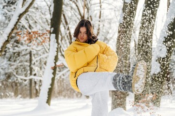 Young female wearing a bright yellow puffer jacket and gray pants is playfully kicking snow in a winter forest setting with snow-covered trees in the background