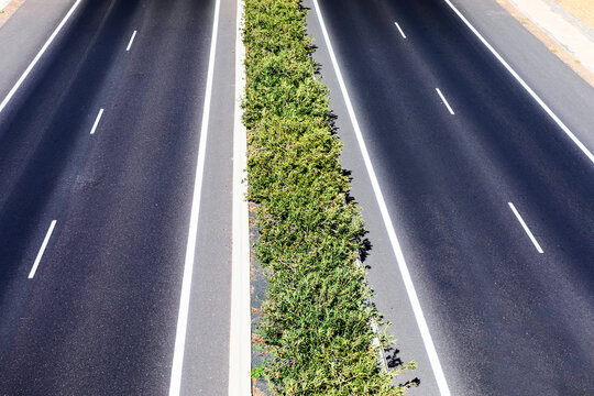 Overhead view of highway separated by a narrow median strip densely planted with green shrubs. Aerial perspective emphasizing geometric pattern of road markings against dark pavement and central hedge
