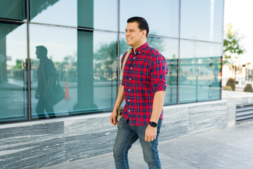 Happy hispanic man walking in urban setting, smiling and carrying bag, wearing plaid shirt and smartwatch
