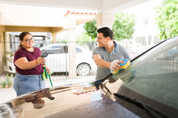 Smiling man and woman cleaning family car with hose and sponge, having fun doing chores in their...