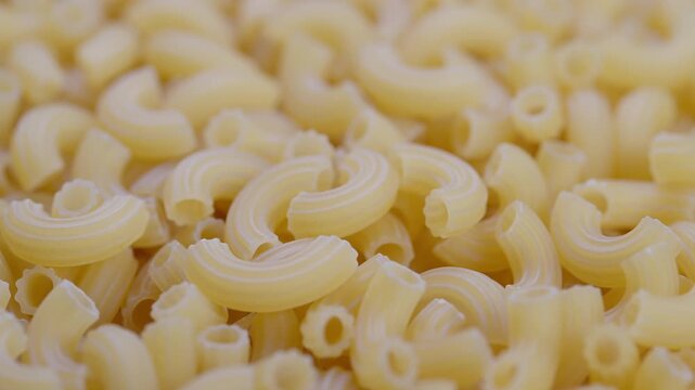 Closeup uncooked elbow macaroni piled loosely, warm beige tones, soft focus, visible ridges and hollow centers, inviting pantry staple texture for recipe or background use