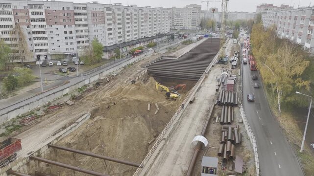 Excavator operates inside deep metro pit surrounded by residential buildings. Heavy machinery works tightly within urban neighborhood