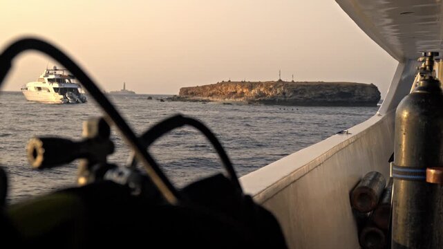 Little Brother Island during sunset. The rocky, arid cliffs of the island are illuminated by the warm, fading light, casting golden tones over the stone and the dark, choppy waters of the Red Sea.