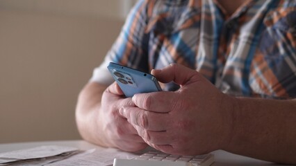 Man using smartphone indoors while sitting at table with keyboard and documents in front of him during daytime