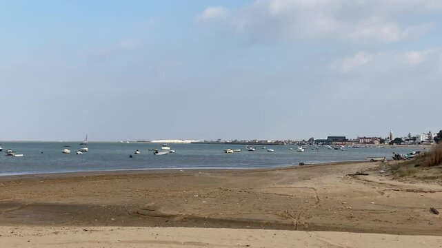 Boats lie at anchor off the sandy shore near Sanlucar, the Bonanza waterfront and lighthouse lining the calm estuary under a soft blue sky