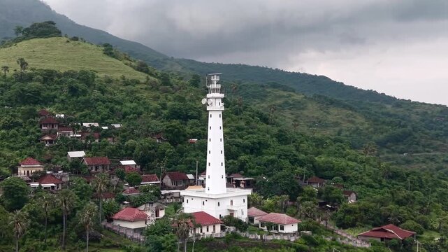 Aerial drone view of white lighthouse on tropical cliff overlooking the ocean, dramatic cloudy sky, coastal landscape in Indonesia.