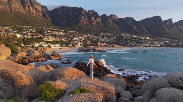 A serene view of Cape Town's coastline at sunset, featuring a woman standing on boulders with Table Mountain in the background
