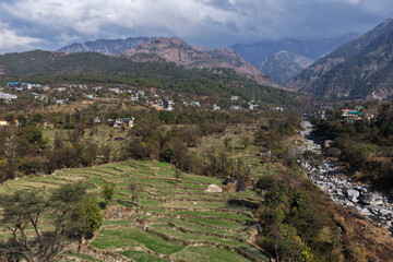An aerial panorama of Dharamshala&rsquo;s lush terraced fields, a rocky riverbed, and the majestic Dhauladhar mountains under a soft, cloudy sky
