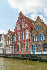 Naklejka premium A row of old brick buildings line a river canal in Bruges. The buildings are of different colors and styles, giving the scene a unique and charming atmosphere. The waterway is calm and peaceful