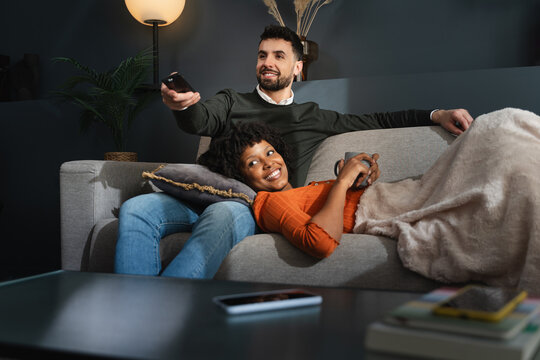Relaxed couple lying on sofa together, watching television online show at night at home