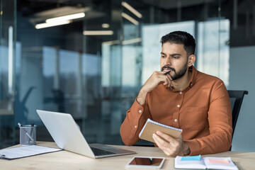 Young man with beard sitting at desk, holding a notebook and pen, looking away from his laptop,...