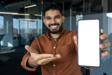 Happy young man presenting a smartphone with an empty white screen for mockups, standing in a contemporary office to highlight mobile technology, communication, and digital services
