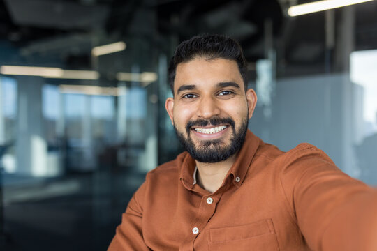 Indian businessman with a beard smiling confidently, taking a selfie in a modern corporate office, representing success, corporate communication, and a positive professional attitude