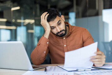 Young male employee feeling tired and frustrated while looking at financial papers at his office...