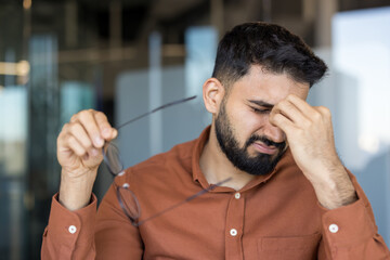 Young man standing in a modern office, experiencing severe eye strain and headache, pinching the...