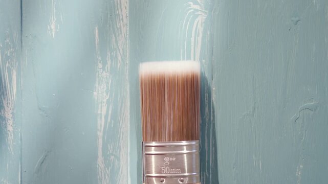 A paintbrush is leaning against a wall. The brush is white and has a brown handle. The wall is blue and has a white paint splatter