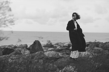 Woman in a long coat stands on rocky coast, wind swept and monochrome, a moody outdoor fashion silhouette by the sea © SHOTPRIME STUDIO