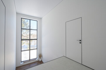 Obraz premium Minimalist room with white walls, large multi-pane window with black frames, sleek white door, light-colored floor, small vase with dried plants near window — airy and bright.