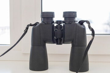 A modern black binoculars with a neck strap stands on a white windowsill against a bright window,...