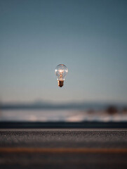 A single glowing light bulb suspended in mid air above a blurred background of road and sky