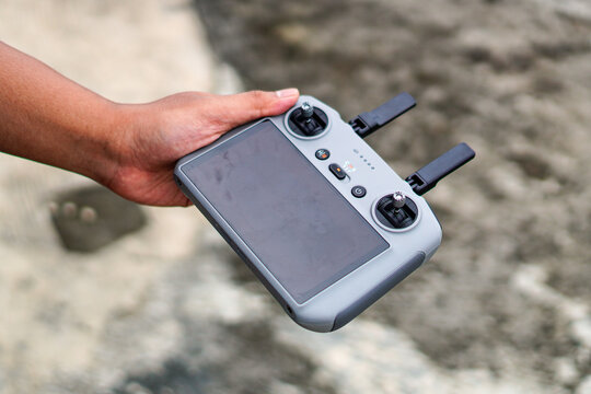 A hand firmly grips a modern drone controller with dual joysticks and antennas against a blurred natural background suggesting outdoor use for aerial photography or videography.