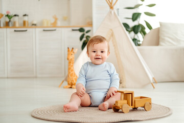 A smiling and joyful kid is playing on the floor with wooden toy cars near the wigwam in the children's room at home, a small child is sitting in a blue bodysuit laughing © Any Grant