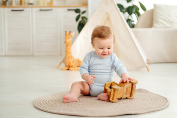 A smiling and joyful kid is playing on the floor with wooden toy cars near the wigwam in the children's room at home, a small child is sitting in a blue bodysuit laughing © Any Grant