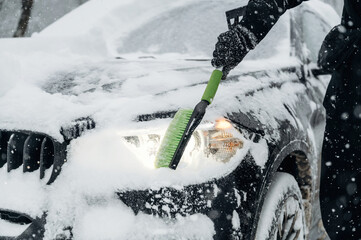 Obraz premium Person using a green snow brush to clear snow from the hood and headlights of a black car during a heavy snowfall in a winter environment with falling snowflakes