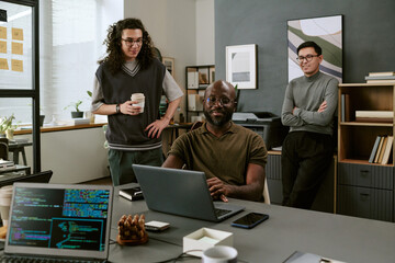 Young Black man sitting at desk using laptop while young adult Caucasian men standing and watching in modern office workspace with computer monitors
