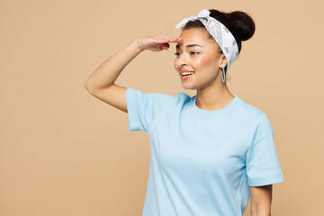 Young woman of African American ethnicity wear blue t-shirt headscarf casual clothes hold hand at forehead look far away distance isolated on plain pastel beige background studio. Lifestyle concept.