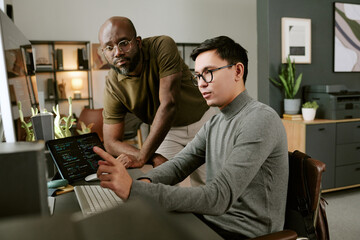 Young Caucasian man sitting at desk coding on computer, while young Black man standing beside him observing screen in modern office workspace with plants and shelves visible
