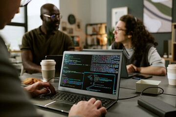 Young adult Caucasian man working on laptop with code on screen, sitting at table with young adult Black man and young adult Caucasian man discussing project in office setting