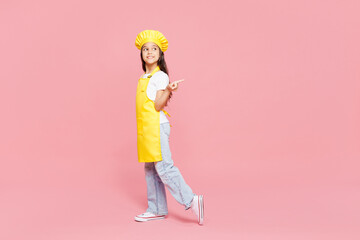 Full body side view small kid child girl housewife chef cook baker mother's helper wears yellow apron toque cap point index finger aside on area isolated on plain pink background Cooking food concept
