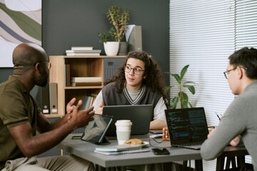 Multiethnic group of young adults collaborating around table, discussing project while using laptops and digital devices, coffee cup and snacks visible, bookshelf in background