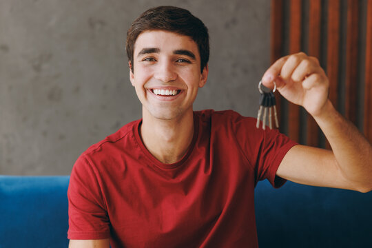 Young happy smiling man he wears red t-shirt hold bunch of keys sit on blue sofa couch stay at home hotel flat rest relax spend free spare time in living room indoors grey wall. People lounge concept.