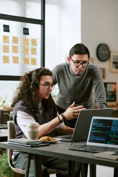 Young adult Caucasian man and young adult Caucasian man collaborating at desk, discussing code on laptop screen, working together on software development project in modern office