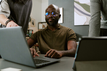 Young Black man sitting at desk using laptop while two colleagues standing nearby collaborating on project in modern office setting, focused on teamwork and technology