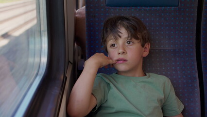 Boy sitting by train window with hand to face, gazing outside with thoughtful expression during travel, candid childhood moment of reflection and stillness