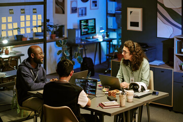 Three young adult men, including Black and Caucasian individuals, collaborating around table with laptops and code on screens, discussing project in modern office workspace