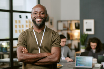 Portrait of young Black man standing with arms crossed and smiling at camera in modern office, two young adult men working on laptops in background, technology workspace setting