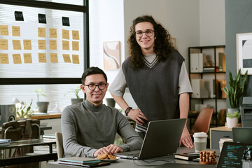 Two young adult Caucasian men working together in modern office, one sitting at desk using laptop while other standing beside him smiling, sticky notes visible on glass wall in background