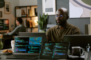 Young Black man working on multiple laptops with programming code on screens in modern office, sitting at desk with coffee cup and snacks, focused on computer tasks