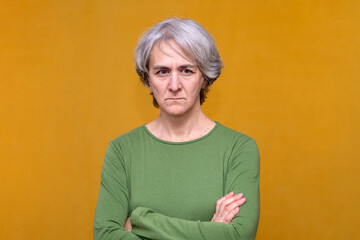 Mature woman with gray hair wearing a green shirt, crossing arms and looking disappointed against a yellow background